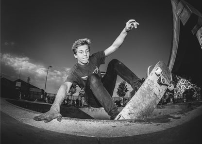 A dynamic, low-angle black and white action shot of a young skateboarder performing a trick in a concrete skatepark. He balances with one hand on the edge of the bowl, his body low to the ground as he carves along the curve, with his other arm raised in the air. Wall Art
