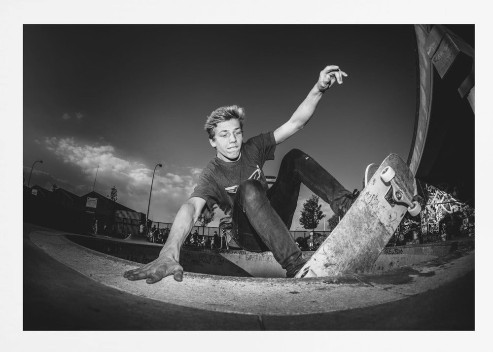 A dynamic, low-angle black and white action shot of a young skateboarder performing a trick in a concrete skatepark. He balances with one hand on the edge of the bowl, his body low to the ground as he carves along the curve, with his other arm raised in the air. Wall Art