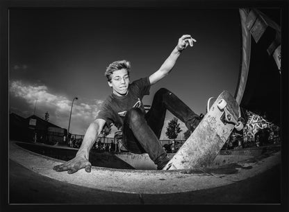 A dynamic, low-angle black and white action shot of a young skateboarder performing a trick in a concrete skatepark. He balances with one hand on the edge of the bowl, his body low to the ground as he carves along the curve, with his other arm raised in the air. Wall Art
