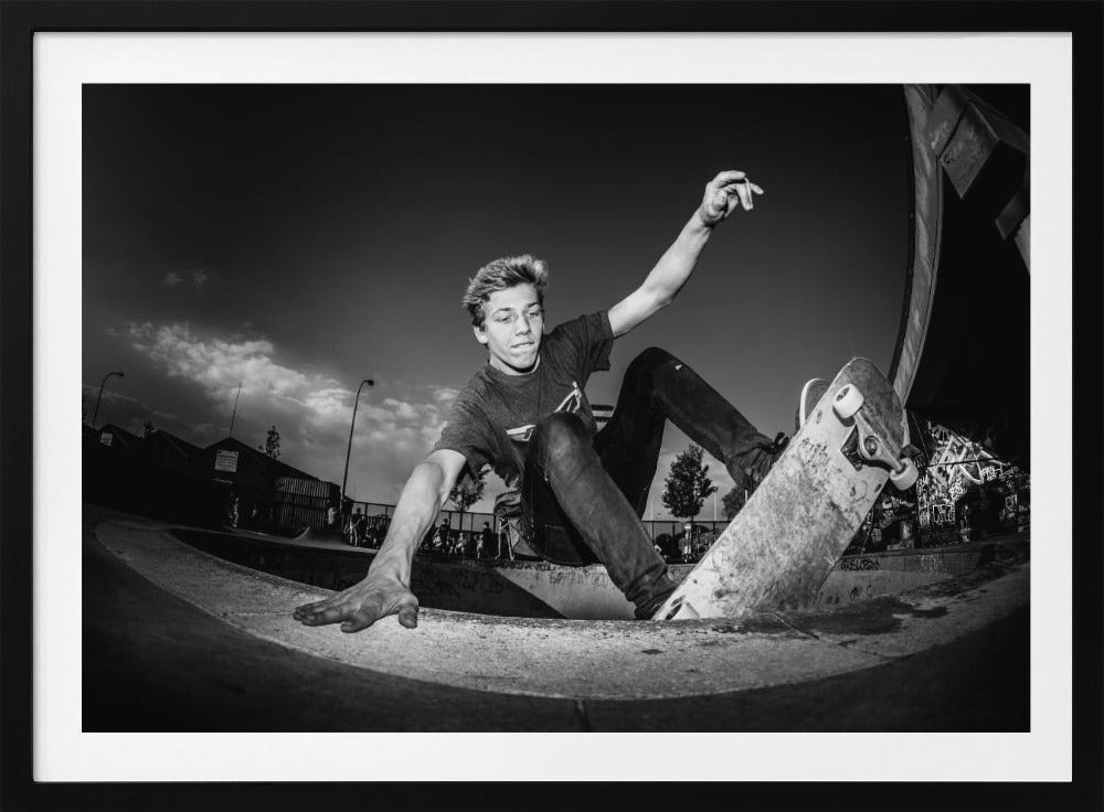 A dynamic, low-angle black and white action shot of a young skateboarder performing a trick in a concrete skatepark. He balances with one hand on the edge of the bowl, his body low to the ground as he carves along the curve, with his other arm raised in the air. Wall Art