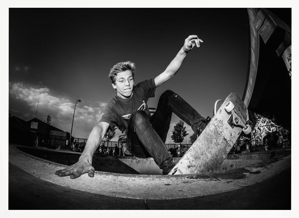 A dynamic, low-angle black and white action shot of a young skateboarder performing a trick in a concrete skatepark. He balances with one hand on the edge of the bowl, his body low to the ground as he carves along the curve, with his other arm raised in the air. Wall Art