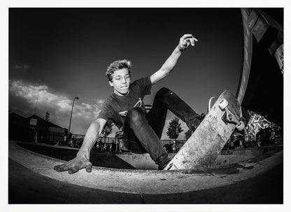 A dynamic, low-angle black and white action shot of a young skateboarder performing a trick in a concrete skatepark. He balances with one hand on the edge of the bowl, his body low to the ground as he carves along the curve, with his other arm raised in the air. Wall Art