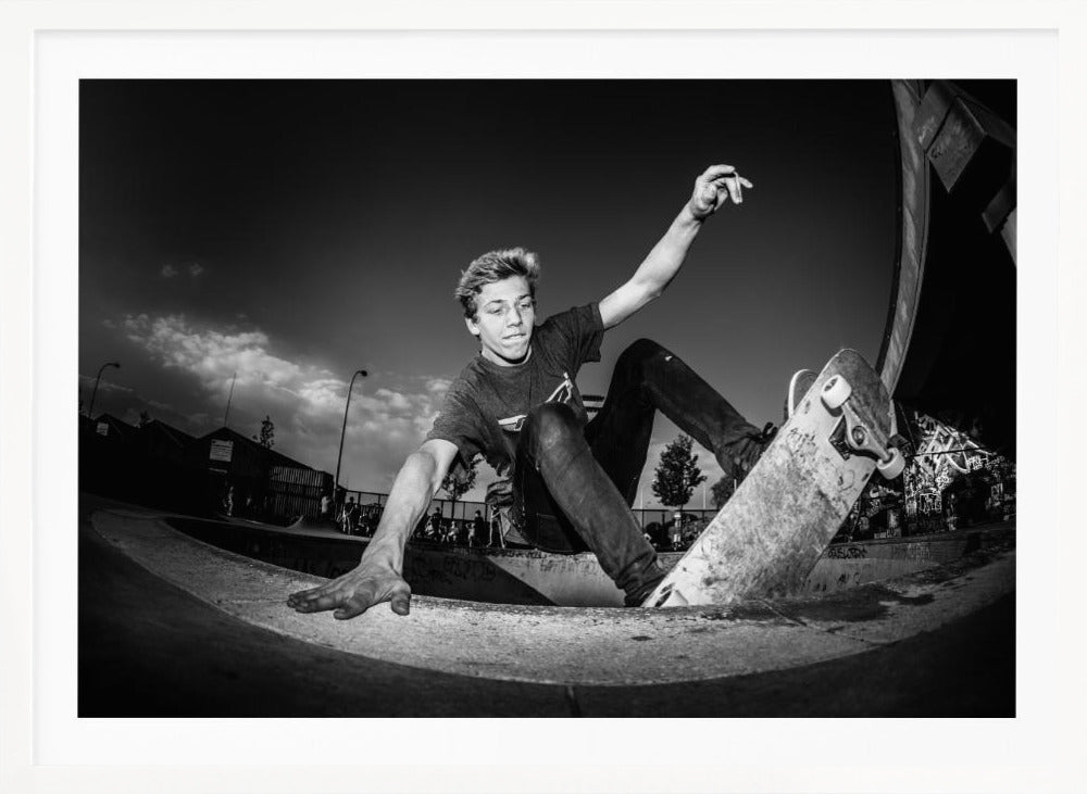 A dynamic, low-angle black and white action shot of a young skateboarder performing a trick in a concrete skatepark. He balances with one hand on the edge of the bowl, his body low to the ground as he carves along the curve, with his other arm raised in the air. Wall Art