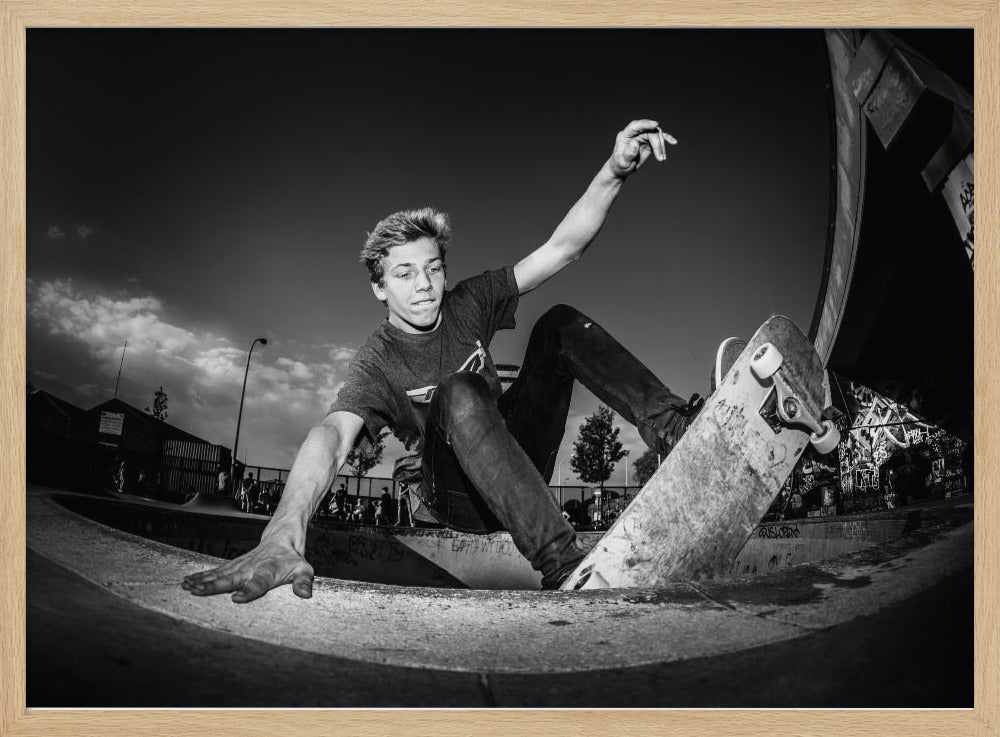A dynamic, low-angle black and white action shot of a young skateboarder performing a trick in a concrete skatepark. He balances with one hand on the edge of the bowl, his body low to the ground as he carves along the curve, with his other arm raised in the air. Wall Art