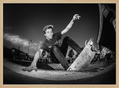 A dynamic, low-angle black and white action shot of a young skateboarder performing a trick in a concrete skatepark. He balances with one hand on the edge of the bowl, his body low to the ground as he carves along the curve, with his other arm raised in the air. Wall Art
