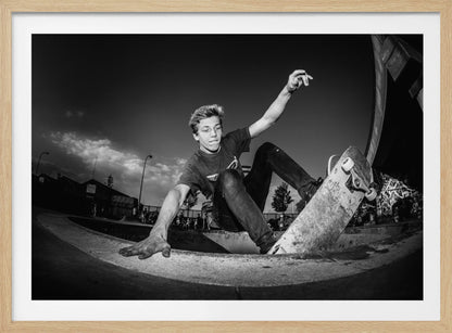 A dynamic, low-angle black and white action shot of a young skateboarder performing a trick in a concrete skatepark. He balances with one hand on the edge of the bowl, his body low to the ground as he carves along the curve, with his other arm raised in the air. Wall Art