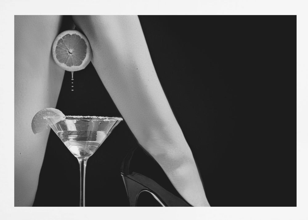 A framed, black and white photograph showing a woman's legs and a martini glass against a black background. A sliced citrus fruit is held between her thighs, with juice dripping towards the sugar-rimmed cocktail glass below. One foot is visible, wearing a black stiletto high heel. Artwork