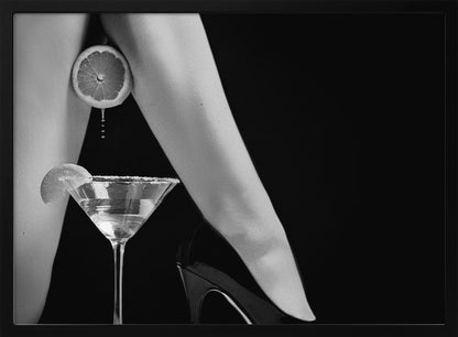A framed, black and white photograph showing a woman's legs and a martini glass against a black background. A sliced citrus fruit is held between her thighs, with juice dripping towards the sugar-rimmed cocktail glass below. One foot is visible, wearing a black stiletto high heel. Artwork