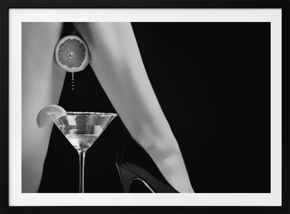 A framed, black and white photograph showing a woman's legs and a martini glass against a black background. A sliced citrus fruit is held between her thighs, with juice dripping towards the sugar-rimmed cocktail glass below. One foot is visible, wearing a black stiletto high heel. Artwork