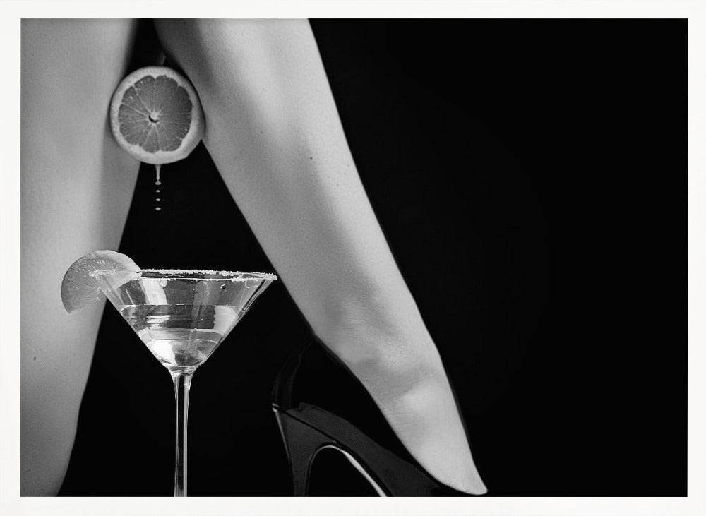 A framed, black and white photograph showing a woman's legs and a martini glass against a black background. A sliced citrus fruit is held between her thighs, with juice dripping towards the sugar-rimmed cocktail glass below. One foot is visible, wearing a black stiletto high heel. Artwork