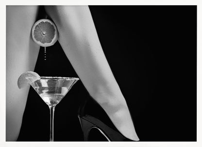 A framed, black and white photograph showing a woman's legs and a martini glass against a black background. A sliced citrus fruit is held between her thighs, with juice dripping towards the sugar-rimmed cocktail glass below. One foot is visible, wearing a black stiletto high heel. Artwork