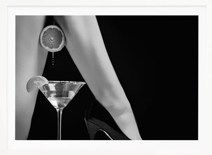 A framed, black and white photograph showing a woman's legs and a martini glass against a black background. A sliced citrus fruit is held between her thighs, with juice dripping towards the sugar-rimmed cocktail glass below. One foot is visible, wearing a black stiletto high heel. Artwork