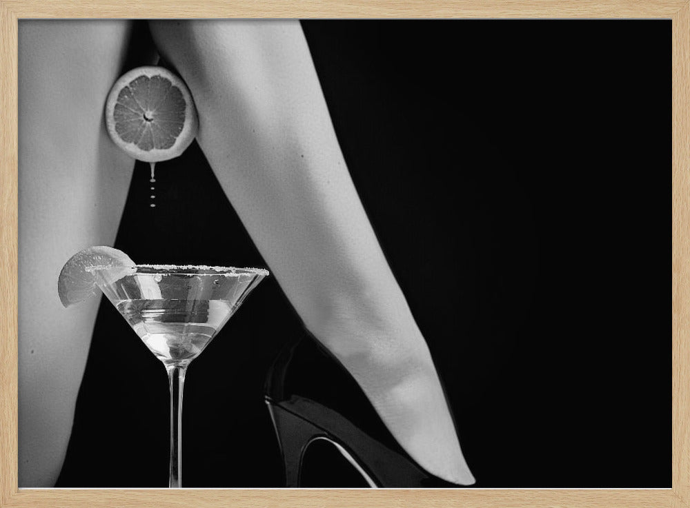 A framed, black and white photograph showing a woman's legs and a martini glass against a black background. A sliced citrus fruit is held between her thighs, with juice dripping towards the sugar-rimmed cocktail glass below. One foot is visible, wearing a black stiletto high heel. Artwork