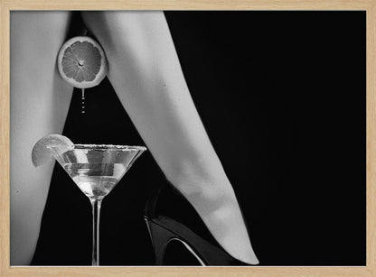 A framed, black and white photograph showing a woman's legs and a martini glass against a black background. A sliced citrus fruit is held between her thighs, with juice dripping towards the sugar-rimmed cocktail glass below. One foot is visible, wearing a black stiletto high heel. Artwork
