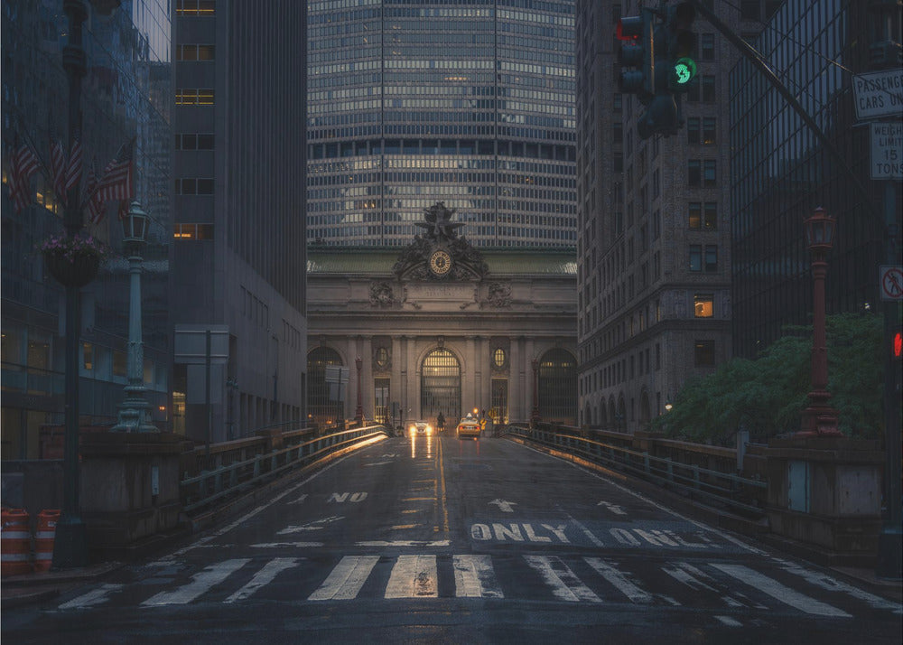 A moody photograph of a wet city street at dusk, leading towards the illuminated facade of Grand Central Terminal in New York City, which is framed by tall, modern skyscrapers. Decor