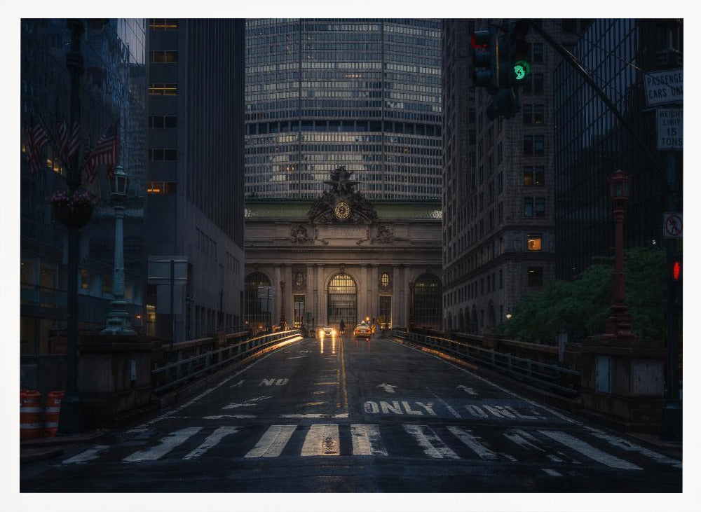 A moody photograph of a wet city street at dusk, leading towards the illuminated facade of Grand Central Terminal in New York City, which is framed by tall, modern skyscrapers. Decor