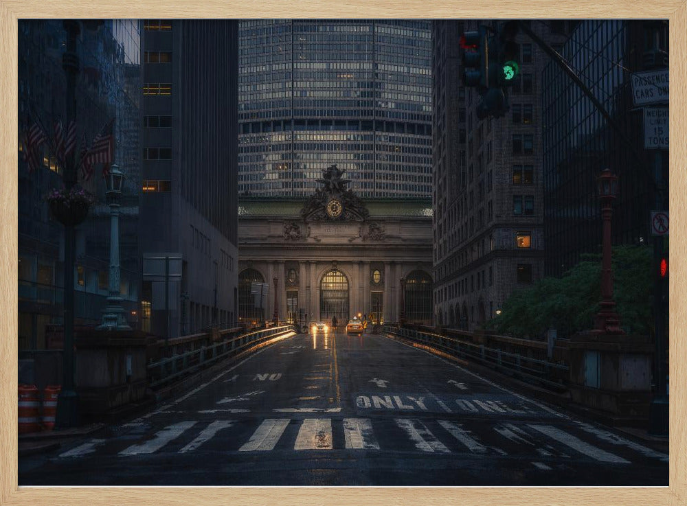 A moody photograph of a wet city street at dusk, leading towards the illuminated facade of Grand Central Terminal in New York City, which is framed by tall, modern skyscrapers. Decor