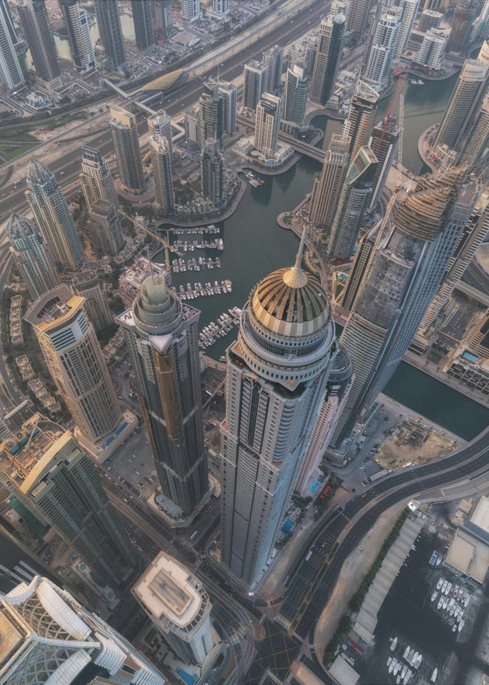 An aerial, bird's-eye view of the dense Dubai Marina, with towering skyscrapers casting long shadows. A waterway snakes through the urban canyon, with numerous yachts docked in the marina. A prominent skyscraper with a distinctive golden dome stands in the foreground. Artwork