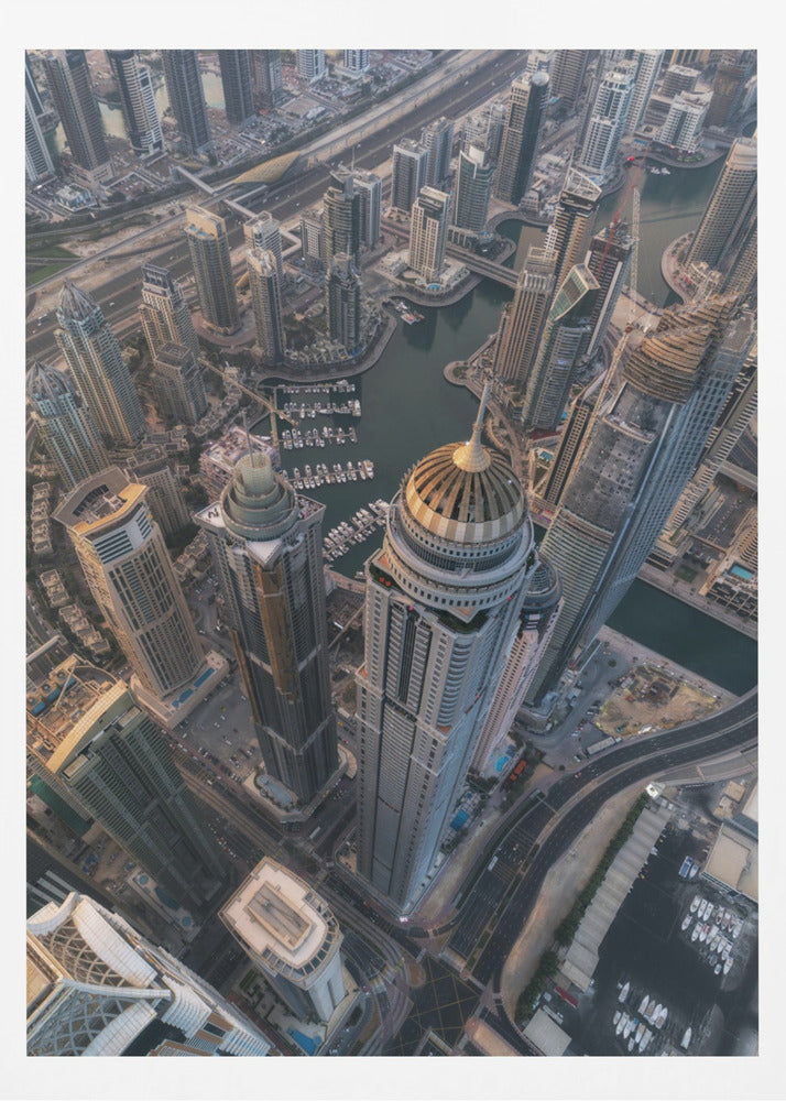 An aerial, bird's-eye view of the dense Dubai Marina, with towering skyscrapers casting long shadows. A waterway snakes through the urban canyon, with numerous yachts docked in the marina. A prominent skyscraper with a distinctive golden dome stands in the foreground. Artwork