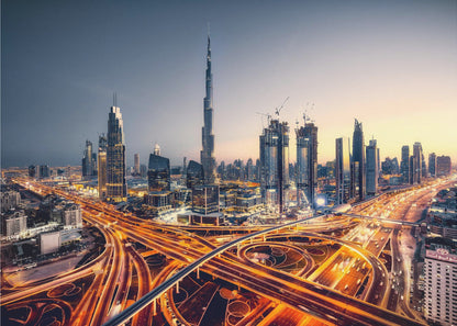 A wide-angle, elevated photograph of the Dubai skyline at dusk, with the iconic Burj Khalifa towering in the center. In the foreground, a complex network of highways glows with the orange light trails of moving traffic, while the city's numerous skyscrapers are illuminated against a twilight sky. Print
