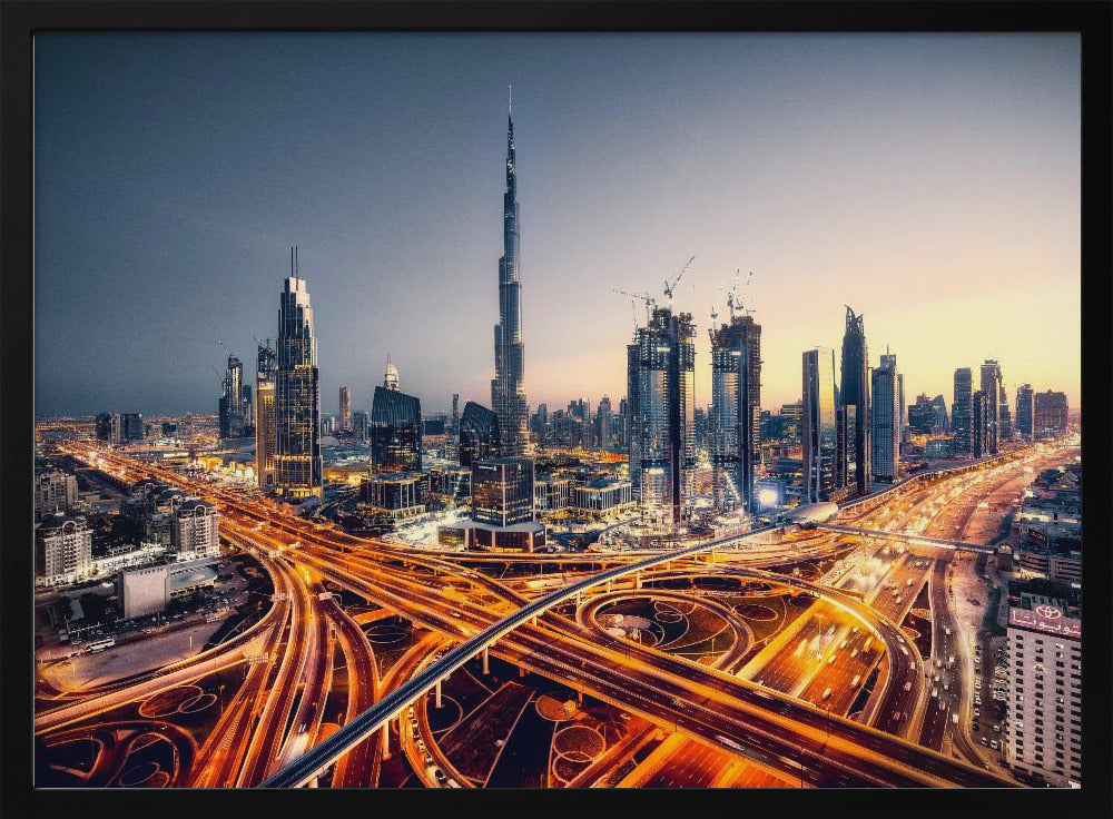 A wide-angle, elevated photograph of the Dubai skyline at dusk, with the iconic Burj Khalifa towering in the center. In the foreground, a complex network of highways glows with the orange light trails of moving traffic, while the city's numerous skyscrapers are illuminated against a twilight sky. Print