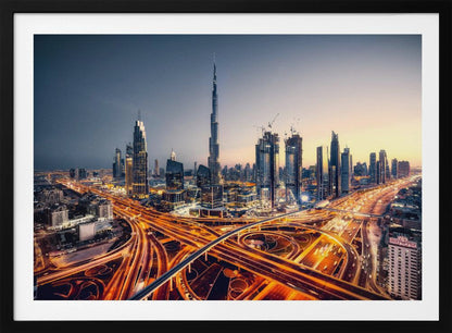 A wide-angle, elevated photograph of the Dubai skyline at dusk, with the iconic Burj Khalifa towering in the center. In the foreground, a complex network of highways glows with the orange light trails of moving traffic, while the city's numerous skyscrapers are illuminated against a twilight sky. Print
