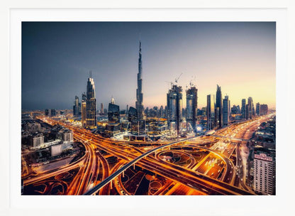 A wide-angle, elevated photograph of the Dubai skyline at dusk, with the iconic Burj Khalifa towering in the center. In the foreground, a complex network of highways glows with the orange light trails of moving traffic, while the city's numerous skyscrapers are illuminated against a twilight sky. Print