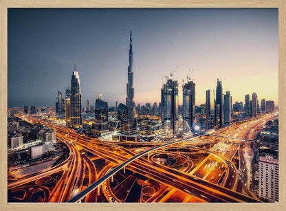 A wide-angle, elevated photograph of the Dubai skyline at dusk, with the iconic Burj Khalifa towering in the center. In the foreground, a complex network of highways glows with the orange light trails of moving traffic, while the city's numerous skyscrapers are illuminated against a twilight sky. Print