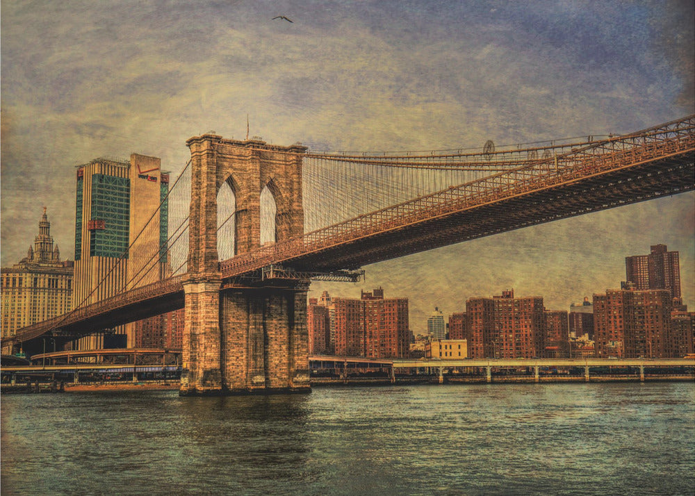 A framed, vintage-style photograph of the Brooklyn Bridge from a low angle, showing its stone pylon and suspension cables against a textured, cloudy sky. The Manhattan skyline and the East River are visible in the background and foreground. Wall Art
