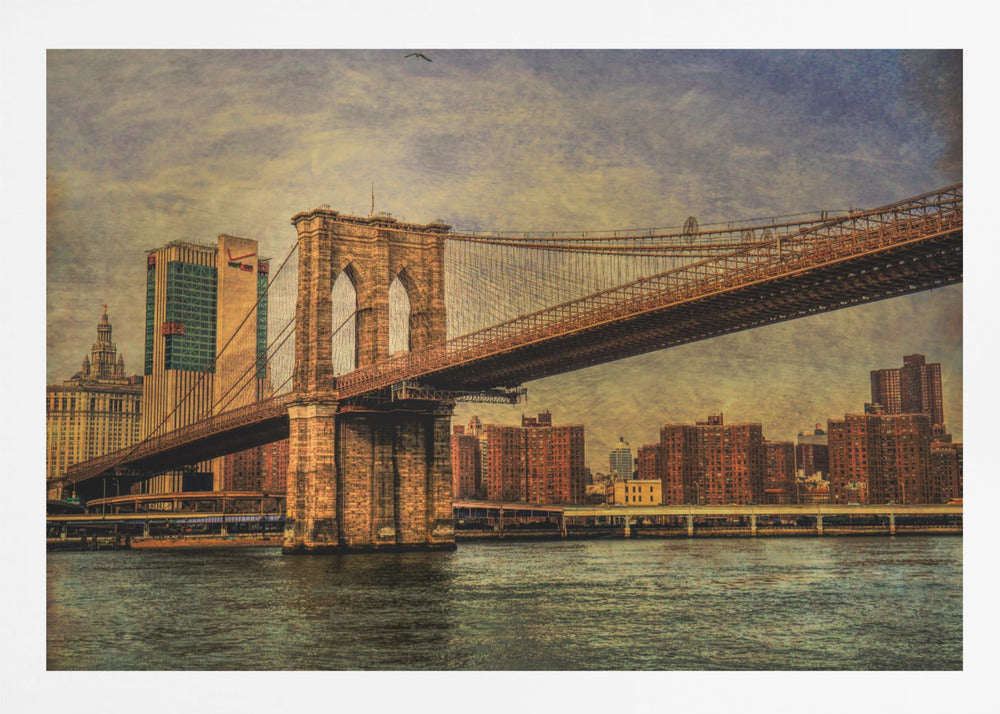 A framed, vintage-style photograph of the Brooklyn Bridge from a low angle, showing its stone pylon and suspension cables against a textured, cloudy sky. The Manhattan skyline and the East River are visible in the background and foreground. Wall Art