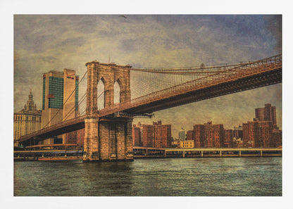 A framed, vintage-style photograph of the Brooklyn Bridge from a low angle, showing its stone pylon and suspension cables against a textured, cloudy sky. The Manhattan skyline and the East River are visible in the background and foreground. Wall Art