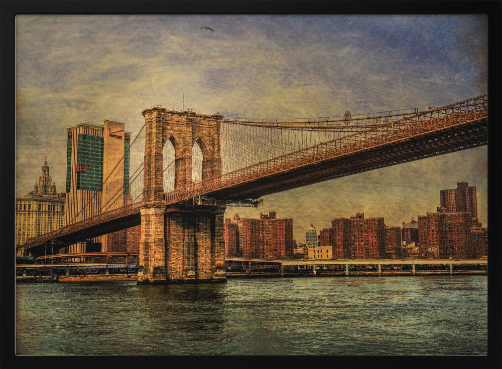 A framed, vintage-style photograph of the Brooklyn Bridge from a low angle, showing its stone pylon and suspension cables against a textured, cloudy sky. The Manhattan skyline and the East River are visible in the background and foreground. Wall Art
