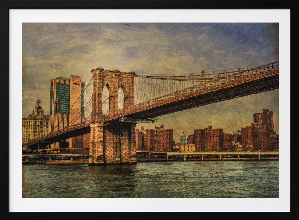 A framed, vintage-style photograph of the Brooklyn Bridge from a low angle, showing its stone pylon and suspension cables against a textured, cloudy sky. The Manhattan skyline and the East River are visible in the background and foreground. Wall Art