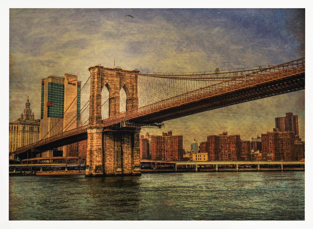 A framed, vintage-style photograph of the Brooklyn Bridge from a low angle, showing its stone pylon and suspension cables against a textured, cloudy sky. The Manhattan skyline and the East River are visible in the background and foreground. Wall Art