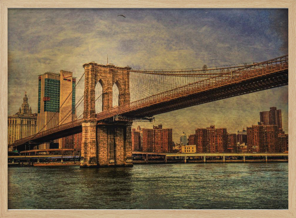 A framed, vintage-style photograph of the Brooklyn Bridge from a low angle, showing its stone pylon and suspension cables against a textured, cloudy sky. The Manhattan skyline and the East River are visible in the background and foreground. Wall Art