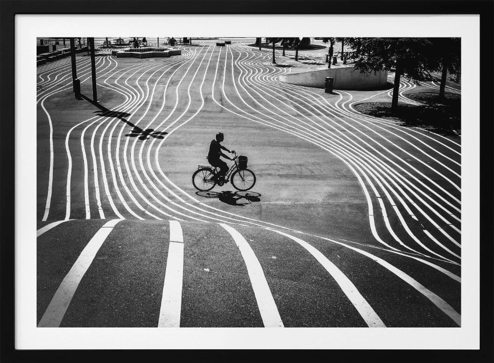 A high-contrast, black and white overhead photograph of a person riding a bicycle across a wide, paved plaza. The ground is covered in a mesmerizing pattern of wavy, parallel white lines, and the cyclist's long shadow is cast by the bright sun. Print