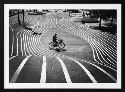 A high-contrast, black and white overhead photograph of a person riding a bicycle across a wide, paved plaza. The ground is covered in a mesmerizing pattern of wavy, parallel white lines, and the cyclist's long shadow is cast by the bright sun. Print