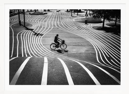 A high-contrast, black and white overhead photograph of a person riding a bicycle across a wide, paved plaza. The ground is covered in a mesmerizing pattern of wavy, parallel white lines, and the cyclist's long shadow is cast by the bright sun. Print