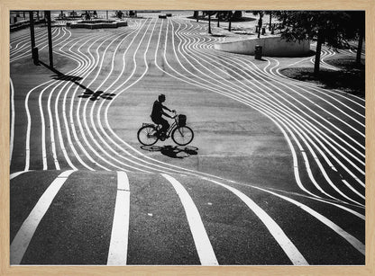 A high-contrast, black and white overhead photograph of a person riding a bicycle across a wide, paved plaza. The ground is covered in a mesmerizing pattern of wavy, parallel white lines, and the cyclist's long shadow is cast by the bright sun. Print