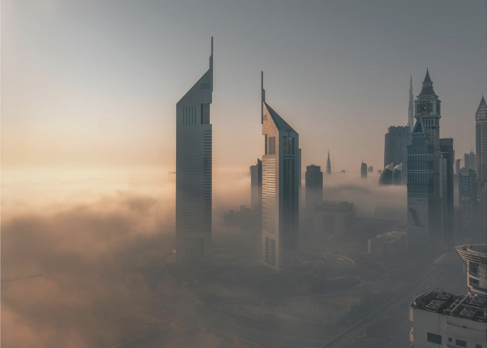 A framed aerial photograph of a futuristic city skyline, likely Dubai, enveloped in a thick layer of fog at sunrise. Tall, modern skyscrapers, including the iconic Emirates Towers, pierce through the clouds, with the warm light of the rising sun creating a hazy, golden glow on the left. Poster