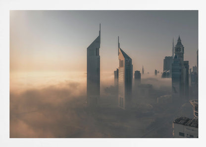 A framed aerial photograph of a futuristic city skyline, likely Dubai, enveloped in a thick layer of fog at sunrise. Tall, modern skyscrapers, including the iconic Emirates Towers, pierce through the clouds, with the warm light of the rising sun creating a hazy, golden glow on the left. Poster