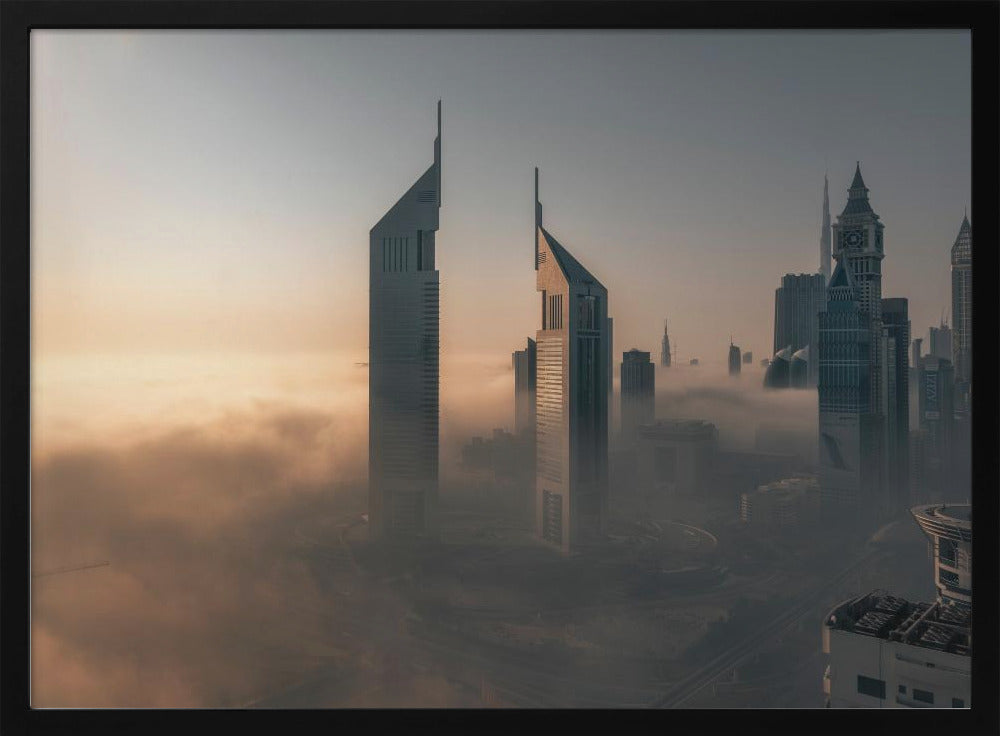 A framed aerial photograph of a futuristic city skyline, likely Dubai, enveloped in a thick layer of fog at sunrise. Tall, modern skyscrapers, including the iconic Emirates Towers, pierce through the clouds, with the warm light of the rising sun creating a hazy, golden glow on the left. Poster