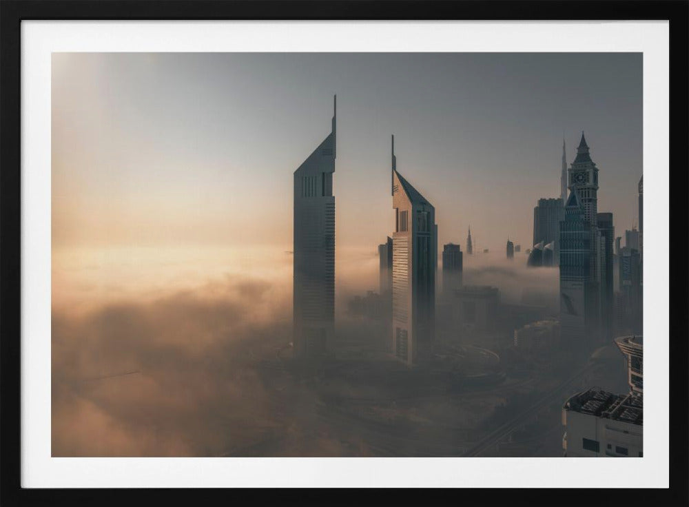 A framed aerial photograph of a futuristic city skyline, likely Dubai, enveloped in a thick layer of fog at sunrise. Tall, modern skyscrapers, including the iconic Emirates Towers, pierce through the clouds, with the warm light of the rising sun creating a hazy, golden glow on the left. Poster