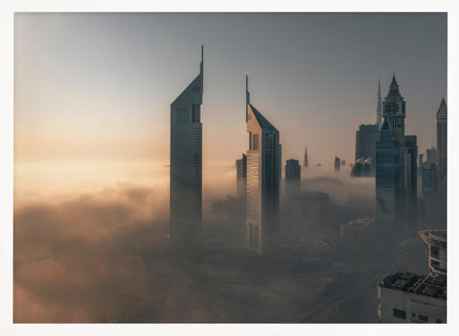 A framed aerial photograph of a futuristic city skyline, likely Dubai, enveloped in a thick layer of fog at sunrise. Tall, modern skyscrapers, including the iconic Emirates Towers, pierce through the clouds, with the warm light of the rising sun creating a hazy, golden glow on the left. Poster