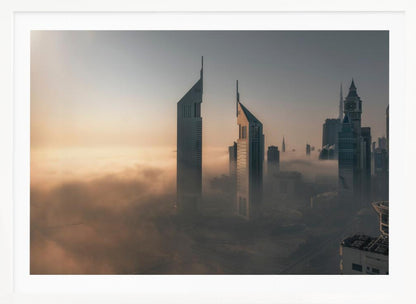 A framed aerial photograph of a futuristic city skyline, likely Dubai, enveloped in a thick layer of fog at sunrise. Tall, modern skyscrapers, including the iconic Emirates Towers, pierce through the clouds, with the warm light of the rising sun creating a hazy, golden glow on the left. Poster