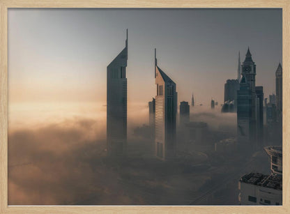 A framed aerial photograph of a futuristic city skyline, likely Dubai, enveloped in a thick layer of fog at sunrise. Tall, modern skyscrapers, including the iconic Emirates Towers, pierce through the clouds, with the warm light of the rising sun creating a hazy, golden glow on the left. Poster