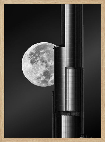 A dramatic black and white photograph of a massive full moon positioned next to a modern, towering skyscraper. The high-contrast image creates a striking juxtaposition between the natural celestial body and the man-made architectural marvel against a dark night sky. Poster