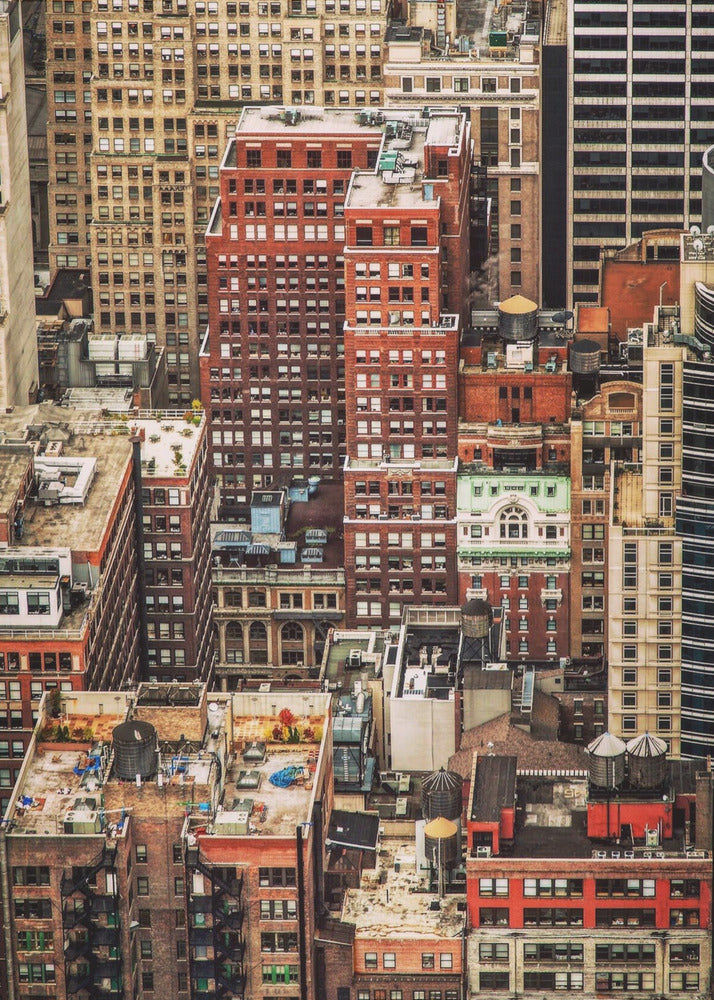 A high-angle, telephoto view of a dense cluster of New York City buildings, featuring a mix of red and brown brick architecture, with numerous windows creating a geometric pattern and several classic wooden water towers perched on the rooftops. Print