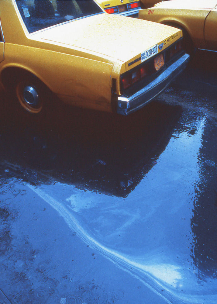 A close-up, angled view of the rear of a yellow taxi cab on a wet street. The car's dark reflection is cast upon a large, vibrant blue puddle in the foreground, with visible ripples from the rain. Wall Art