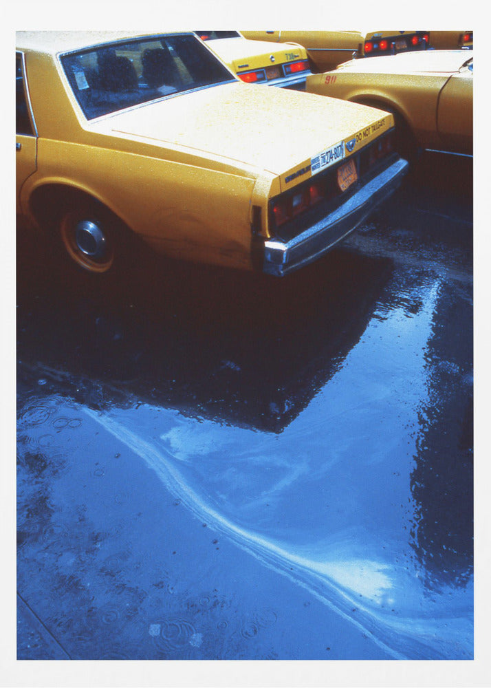 A close-up, angled view of the rear of a yellow taxi cab on a wet street. The car's dark reflection is cast upon a large, vibrant blue puddle in the foreground, with visible ripples from the rain. Wall Art